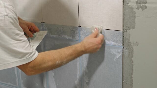 Worker using a rubber grout float to smooth grout between tiles. Carefully applying sealant to tiles joints. Workman hands smoothing caulk, using small tool to remove excess and create clean seam.