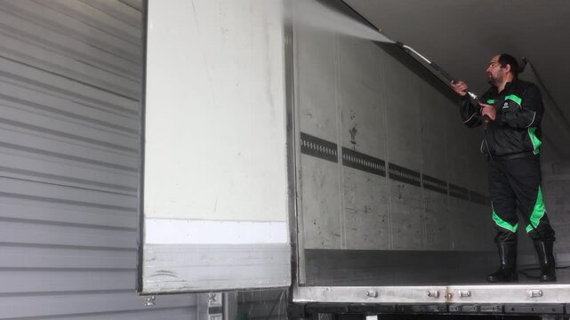 Water sprays reflect sunlight, highlighting the meticulous effort put into making the truck shine like new. A dedicated professional truck cleaner thoroughly washing a semi-trailer truck.