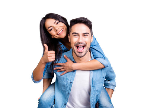 excited two people in love is having fun, they adore each other. Pretty joyful lovely smiling woman is sitting on her boyfriend's back and showing thumb up, they are isolated on grey background