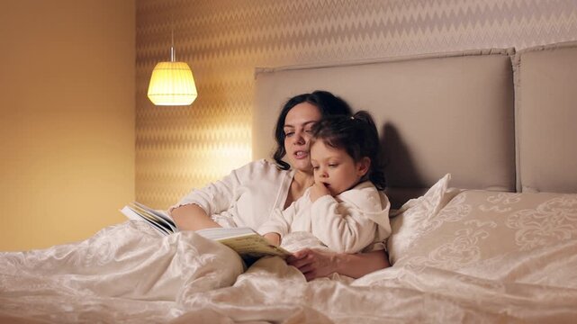 Loving mother reading a book to her little daughter in bed before sleep. A cozy family moment sharing a bedtime story in the evening