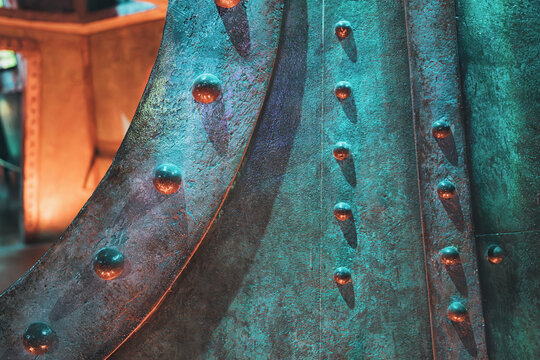 Detailed view of a textured industrial metal surface, showing patinated bronze and copper colors with round rivets in green and orange light