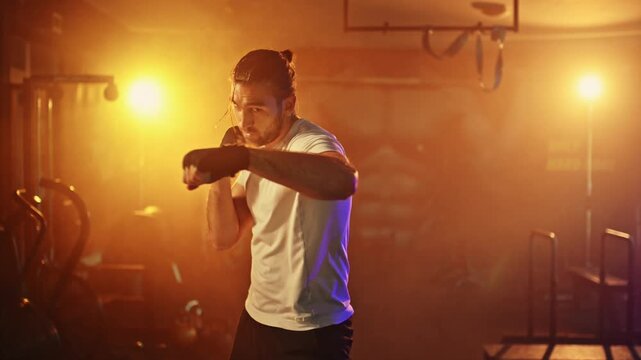 Determined male boxer shadowboxing in a dark gym with dramatic lighting. Muscular athlete training martial arts and fighting skills