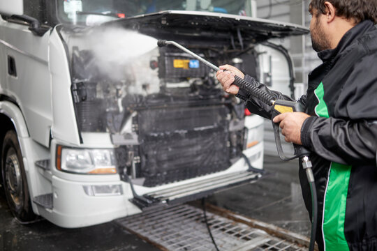 This image captures a professional cleaner utilizing a contactless washer to clean beneath the hood of a white truck in a large car wash hangar, showcasing effective vehicle maintenance techniques.