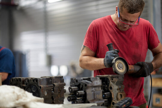 A diligent service technician wearing safety glasses meticulously cleans a cylinder head inside a well-equipped truck garage. The background features various automotive tools and equipment.