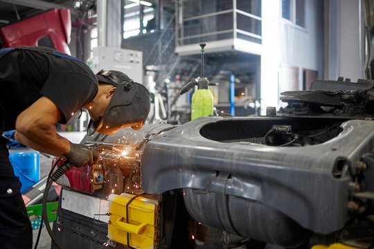 .A dedicated service center employee, wearing a protective mask, skillfully welds to repair a truck. The scene captures the precision and attention to detail involved in vehicle maintenance.