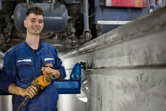 A joyful mechanic dressed in blue overalls passionately repairs a truck's suspension using a pneumatic tool in a well-lit service pit, showcasing expertise and dedication in automotive maintenance.
