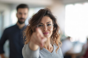 Obraz premium Woman pointing finger toward camera with confident expression while coworker stands in background slightly blurred, bright office interior, natural window light isolated in contemporary studio setup