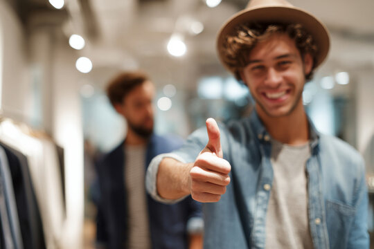 Close up of young man giving thumbs up while shopping with friend in clothing store, modern retail interior softly blurred, bright store lighting, casual lifestyle moment with neutral tones