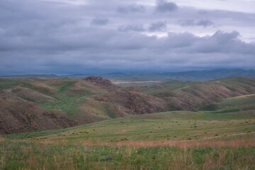 Obraz premium Panoramic view of rolling green hills and mountain ridges under a cloudy sky in Kazakhstan. Natural landscape background suitable for travel, environment, and outdoor adventure 