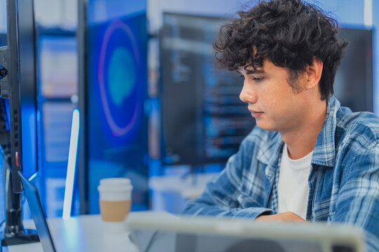 focused young Asian male programmer with curly hair is working intently on his laptop in a modern data center office. The background features bright blue neon LED-lit monitor
