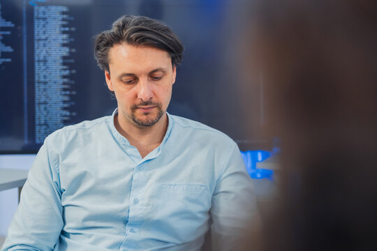 A confident male tech professional or business consultant in a light blue shirt engaged in a collaborative discussion with a colleague. They are sitting in a high-tech office environment with large ba