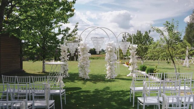 Beautiful outdoor wedding ceremony area with chairs for guests. Wedding arch decorated with flowers and glass beads in summer outdoors. Decorated wedding ceremony venue. Camera pull back.