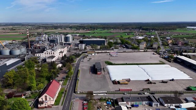 Aerial view of a sprawling agricultural processing and storage industrial complex on a sunny day, featuring grain silos, a smokestack, production buildings, a large parking lot, white silage storage b