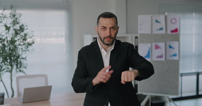 Portrait of Middle Eastern businessman threatens with finger and points at wristwatch in office. Serious male manager reminds about missed deadline in boardroom