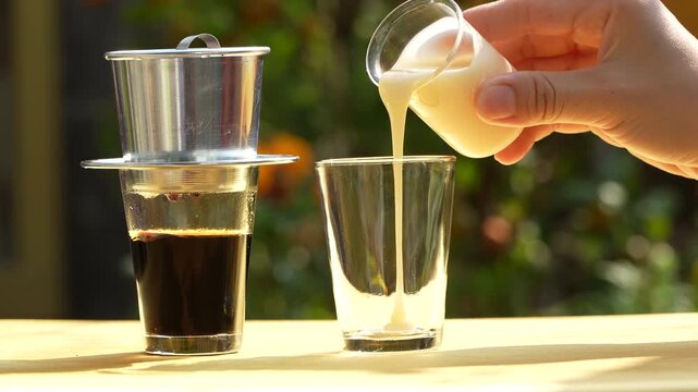 Pouring condensed milk for traditional Vietnamese iced coffee with phin filter in sunlight.