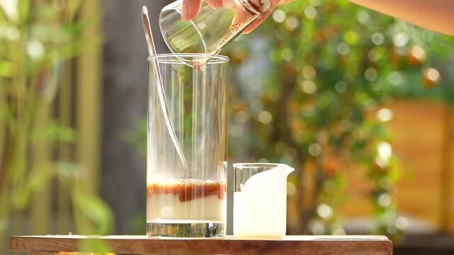 Pouring coffee into condensed milk in a tall glass under golden morning sunlight.