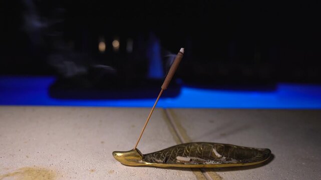 Burning incense stick on golden leaf holder with smoke at night outdoors near swimming pool with blue lighting meditation and aromatherapy concept for spa wellness and relaxation