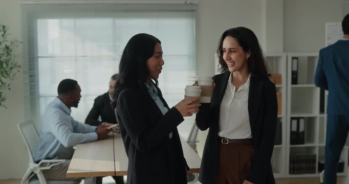 Portrait of Asian and Latin business women smile and share coffee during project talk in company office. Colleagues raise cups and discuss tasks during break