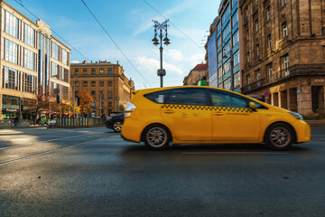 Fototapeta premium Yellow taxi cab driving fast on a city street in Budapest Hungary with blurred motion effect and beautiful European architecture under a bright blue sky during daytime