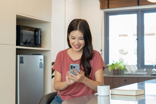 Smiling Asian Woman Using Smartphone with Coffee in Kitchen