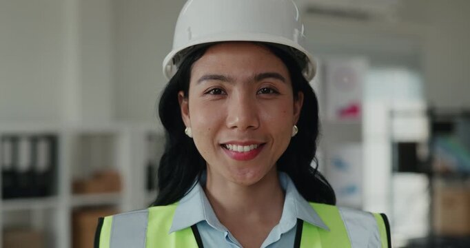 Portrait of Asian engineer smiles in hard hat and safety vest in office. Specialist looks at camera and stands confidently during construction planning session