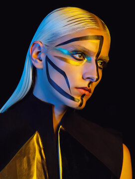 Avant-garde portrait of a woman with slicked-back platinum hair and striking geometric face paint. Bold black lines and metallic gold accents create a futuristic, high-fashion aesthetic