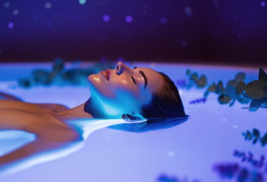 Woman floats peacefully in a glowing wellness bath, surrounded by floating eucalyptus and lavender plants. Ethereal blue water creates a dreamy atmosphere of relaxation, healing and sensory escape