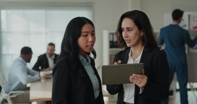 Portrait of Asian and Latin business women look at tablet in office as team works near planning board. Colleagues hold screen steady and compare project notes