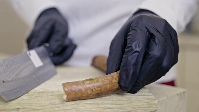 A culinary expert, wearing black gloves, precisely cuts a savory sausage into bite-sized pieces on a wooden cutting board, showcasing food preparation with focus and hygiene.