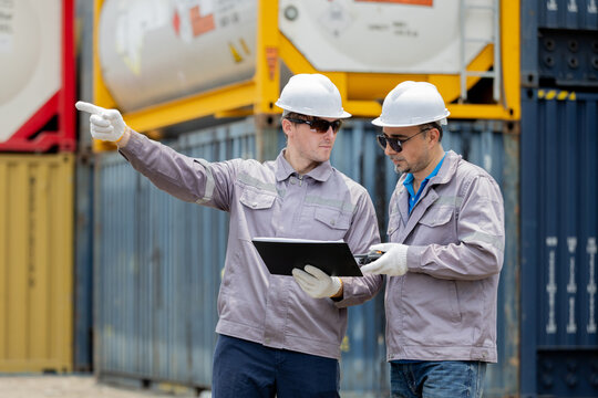 Two workers in safety gear discuss logistics at a shipping container yard, with one pointing and the other checking a tablet.