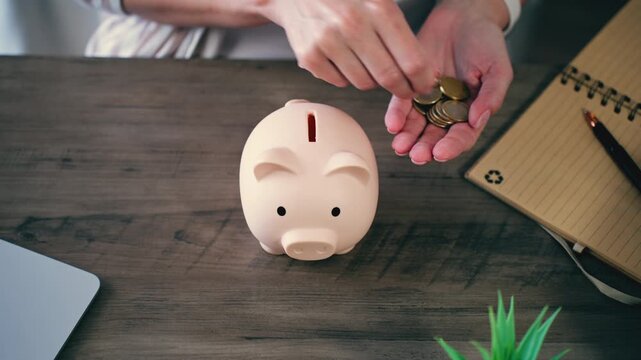 Woman sitting at wooden desk inserting euro coins into pink piggy bank, illustrating concept of saving money, personal finance management, investment, and planning for retirement