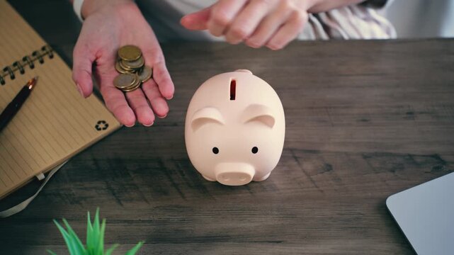 Woman&rsquo;s hands insert coins into a pink piggy bank on wooden desk, symbolizing financial planning, savings accumulation, investment strategy and future financial security concept at home.