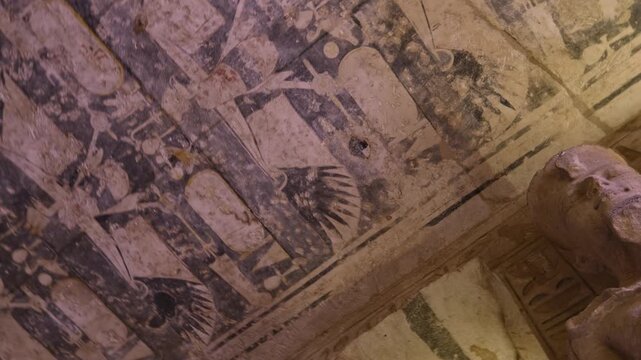 Close-up of faded ancient ceiling inside Abu Simbel temple, Egypt, with deteriorated hieroglyphics, winged vultures, cartouches and sacred symbols on aged stone surface.