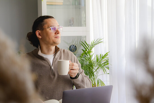 Middle aged man enjoying coffee break while working remotely near bright window. Peaceful pause, work life balance, productivity and calm home office routine concept.