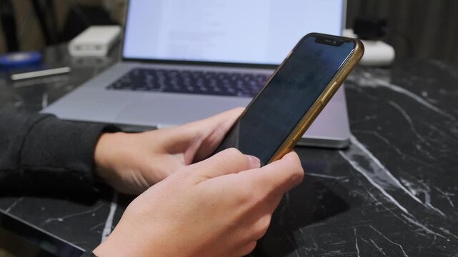 Close-up of hands deleting text on smartphone screen while sitting at desk with laptop, showing mobile typing correction, messaging workflow, digital communication, everyday device usage