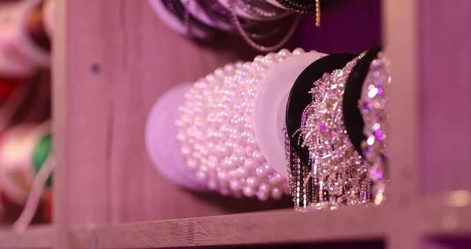 Close up of a hand gently holding a sparkling silver rhinestone ribbon inside a haberdashery store. Wooden shelves filled with various colorful trims are blurred in the background.

