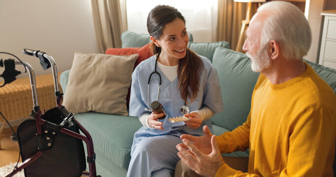 A home health nurse talks to an elderly man in a living room. The nurse holds a bottle and smiles while the man sits comfortably. A walker is nearby, indicating support and assistance.