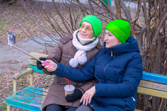 Daughter and senior mother taking selfie on smartphone with monopod while sitting on park bench. Happy family lifestyle outdoors during cold autumn day. Connection and technology