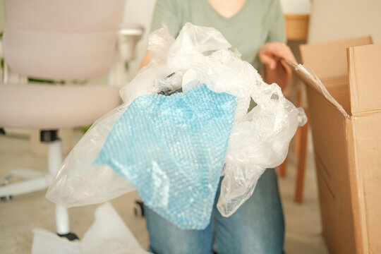 A person holding a bundle of plastic waste, including bubble wrap and bags, in a living room setting. A cardboard box is nearby, indicating unpacking.