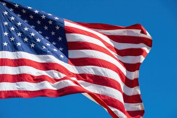 Fototapeta premium Vibrant American flag proudly waves against a brilliant blue sky, showcasing the iconic red stripes and white stars, symbolizing freedom, patriotism, and national pride in the USA.
