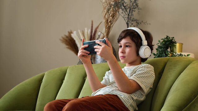Side portrait of a boy in headphones holding a phone as he strokes a beagle in a cozy living room. Mobile media, pet companionship, and family downtime reflect modern childhood at home.
