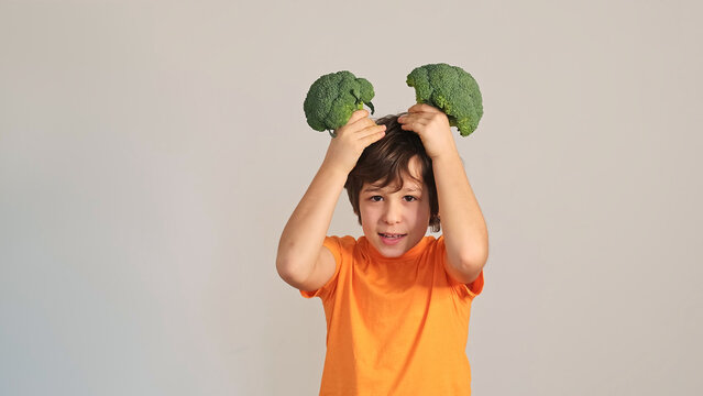 Front view of a boy in an orange shirt holding two broccoli heads above his hair like playful horns against a plain wall. Healthy food, picky eating, and family meals shape childhood nutrition habits.