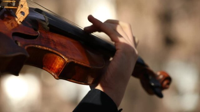 A close up of skilled hands playing a classic wooden violin outdoors. The musician gracefully moves the bow across strings, creating beautiful melodies against a blurred natural background.

