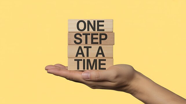 Hand holding stacked wooden blocks with "one step at a time" small steps patience progress methodical motivation inspiration goal setting achievement persistence endurance deliberate