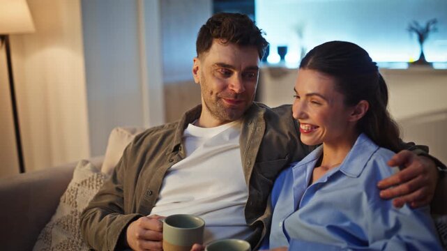 Happy lovers embracing sofa drinking evening coffee together. Man cuddling woman