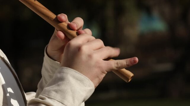 A close-up shot of a musician's hands skillfully playing a traditional wooden flute outdoors. Natural sunlight highlights the instrument and delicate finger movements against a beautifully blurred bac