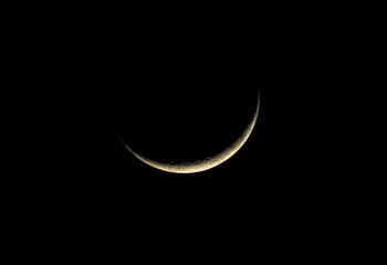 A sharp waxing crescent moon hangs in the pitch-black night sky. This high-contrast shot reveals the rugged texture of craters along the lunar edge, glowing with a soft, warm golden light. © daniel