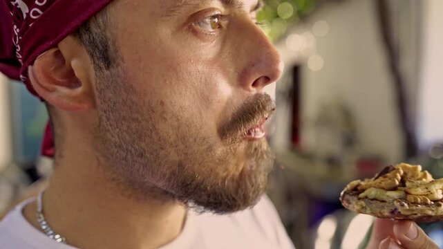 A bearded man in a red bandana savors a delightful, homemade cookie, topped with crunchy nuts and sweet ingredients. He takes a bite, visibly enjoying the healthy, tasty treat as an outdoor snack.