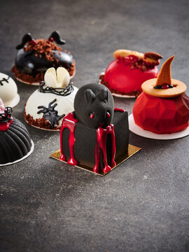 The image showcases an assortment of unique Halloween-themed desserts, including a cat-shaped cake with red glaze