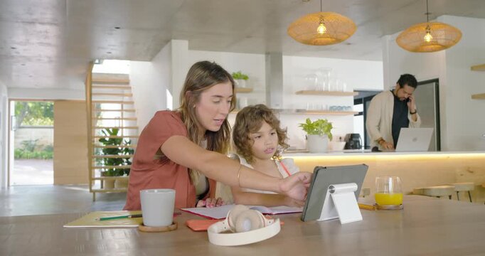 Family mother and daughter pointing at tablet on stand, guiding schoolwork while father calling